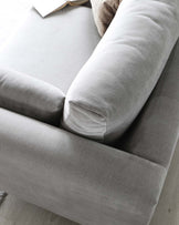 Contemporary light grey fabric sofa with plush cushions and a cylindrical bolster pillow, featuring a sleek and comfortable design with a soft texture, perfect for modern living spaces; a chic throw and an open book rest casually on the sofa, adding a touch of lived-in elegance to the setting.