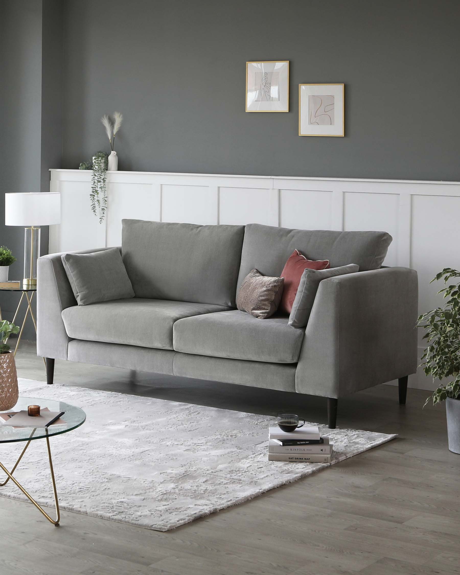 Contemporary three-seater sofa upholstered in a light grey fabric with plush cushions and clean lines, standing on minimalist black legs. A white textured area rug sits underneath, with a round glass-top coffee table featuring gold metal legs to the side. A sleek white table lamp with a gold base complements the elegant decor.