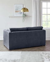 Modern charcoal grey velvet loveseat with sleek lines and minimalistic design, featuring plush back cushions and a low-profile silhouette, resting on subtle dark wooden legs. Positioned on a textured light grey and white area rug in a well-lit room with neutral decor.