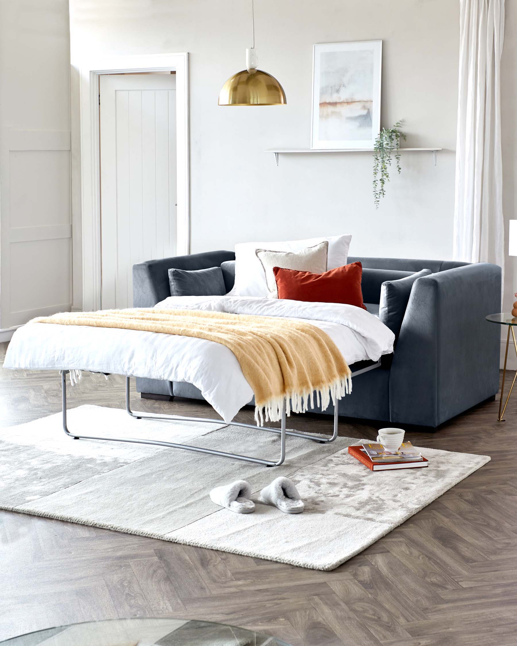 Modern minimalist bedroom featuring a sleek, low-profile platform bed with a white bedspread and a caramel-coloured throw blanket, complemented by a deep blue upholstered sofa with matching and contrasting pillows. A contemporary, chrome-framed bench sits at the foot of the bed, while a plush, neutral-toned area rug anchors the space. A small gold and glass side table and a modern gold pendant light add metallic accents, enhancing the luxurious yet understated ambiance.