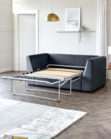 Modern convertible sofa bed in dark grey upholstery with metal frame, extended into bed position with wooden slats visible. A small round side table with golden legs and a marble top is placed to the right. The room features contemporary decor with minimal wall art, a metallic pendant light, and light coloured area rugs on a herringbone parquet floor.