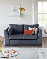 A contemporary charcoal grey upholstered sofa with plush cushions, accented by a terracotta and two beige throw pillows. In front, a round glass coffee table with a golden geometric frame holding books, situated atop an area rug with a mix of grey and off-white tones. To the right, a modern side table with a white top and golden legs, featuring a small white lamp.