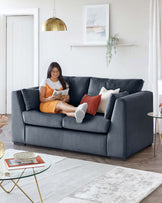 A contemporary charcoal grey fabric three-seater sofa with plush cushions and a structured silhouette, paired with a round glass-top coffee table featuring a gold geometric base. A white and grey area rug underlines the set.