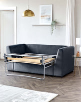 A modern charcoal grey sofa bed with a visible metallic pull-out frame and wooden slats partially extended, set in a contemporary living room with light wood flooring, a small round side table with a gold base and a white top, a simple white area rug, and minimalist decor.