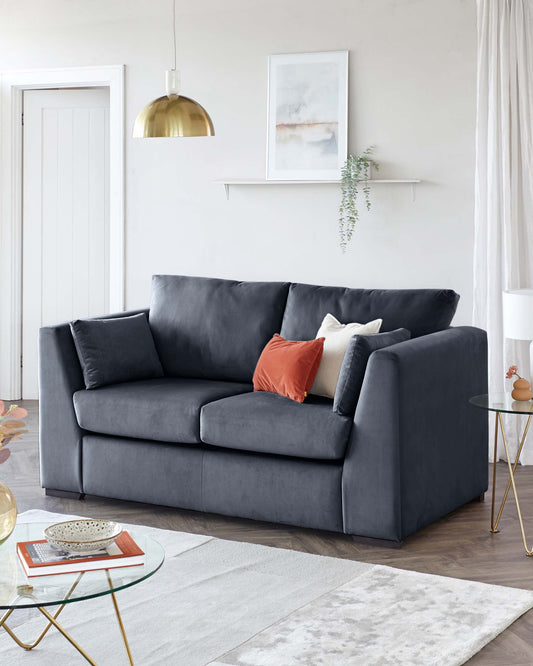 A contemporary charcoal grey upholstered sofa with plush cushions and a clean-lined silhouette. In front of the sofa is a modern round glass-top coffee table with a gold geometric frame. The room is accented with a white and grey patterned area rug.