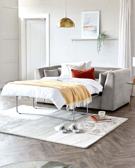 Modern living room with a light grey upholstered sofa with plush cushions, a minimalistic metal frame coffee table with a glass top, and a round side table with a golden finish. The scene is completed by a large, soft area rug in muted tones, set on a herringbone-patterned wooden floor. A chic, golden pendant light hangs above, adding a warm glow to the space.