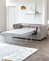 A contemporary light grey corner sofa with a convertible bed function, partially extended to showcase the metal frame and mattress. The room also features a white area rug with a subtle pattern and a small round side table with a gold frame and dark tabletop.