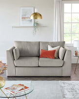 A contemporary three-seater sofa upholstered in light grey fabric with plush back cushions and one bright red accent pillow. In front of the sofa is a round glass-top coffee table with a geometric gold metal base. The room is complemented by a soft grey area rug beneath the coffee table.