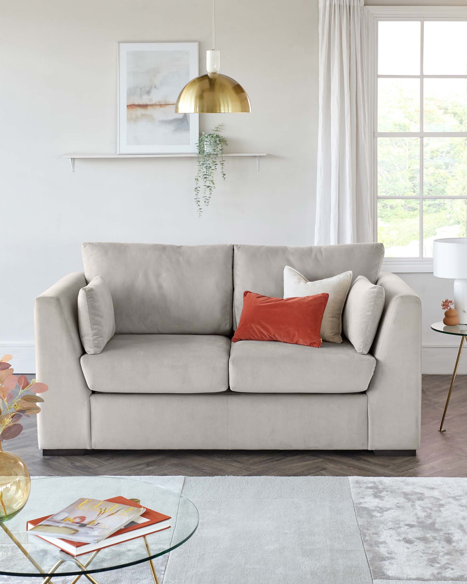 Contemporary light grey upholstered sofa with plush cushions and a mix of matching and contrasting throw pillows. In front of the sofa is a modern round glass-top coffee table with a gold geometric base. A textured light grey area rug sits underneath the furniture pieces.