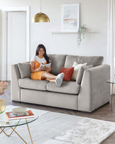 A contemporary living room scene featuring a light grey upholstered three-seater sofa with cosy cushioned arms and back, complemented by white and rust throw pillows. In front of the sofa is a round glass top coffee table with a gold geometric metal base. Underneath is a textured grey and white area rug with an abstract pattern. The room is accented with a gold dome pendant light and a floating white shelf with a hanging green plant.