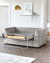 Modern light grey upholstered sofa bed with a pull-out metal frame and wooden slats, partially extended to showcase the convertible mechanism, in a contemporary living room setting with minimalist decor. A small round side table with a glass top and gold-metal legs is beside the couch.