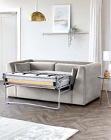 Modern light grey fabric sofa bed with a pull-out metal mechanism on display, accompanied by a small round side table with a glass top and gold-finished metal legs. The room has a minimalist style with a white interior, wooden flooring, and subtle decorative elements.