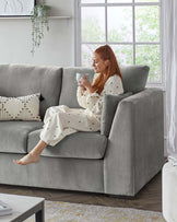 A modern light grey upholstered three-seat sofa with plush cushions and structured armrests, situated in a cosy bright room with sheer curtains and natural light.