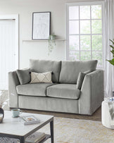 A contemporary living room featuring a comfortable, plush light grey fabric sofa with three seat cushions and backrests, accompanied by a patterned throw pillow. In front of the sofa is a modern rectangular coffee table with a white marble top and a black metal frame. The room also has a beige and yellow patterned area rug beneath the furniture.