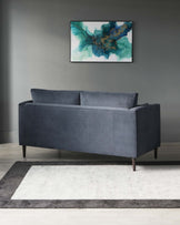 A modern charcoal grey fabric two-seater sofa with a clean-line silhouette and minimalistic dark wooden legs. Positioned on a two-toned grey and white area rug with a distinctly contemporary geometric design. The wall behind features a tranquil abstract painting with blue and green hues, which complements the clean and sophisticated aesthetic of the space.