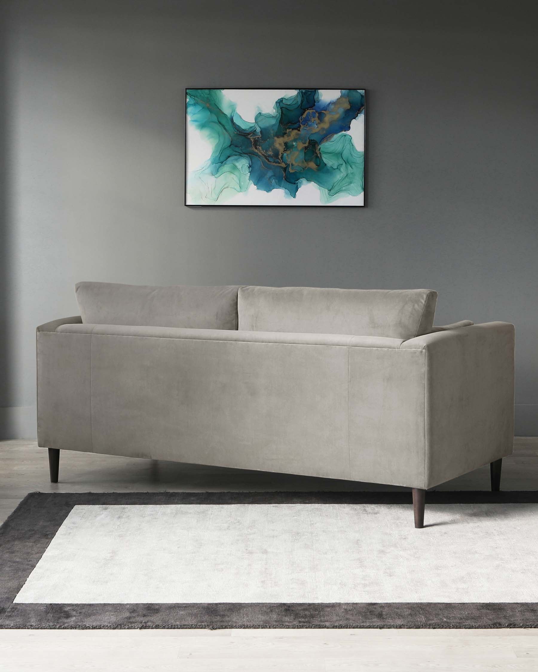 A modern light grey velvet sofa with clean lines and dark wooden legs, positioned on a two-tone grey area rug against a grey wall. A contemporary abstract art piece with blue and green hues hangs above the sofa.