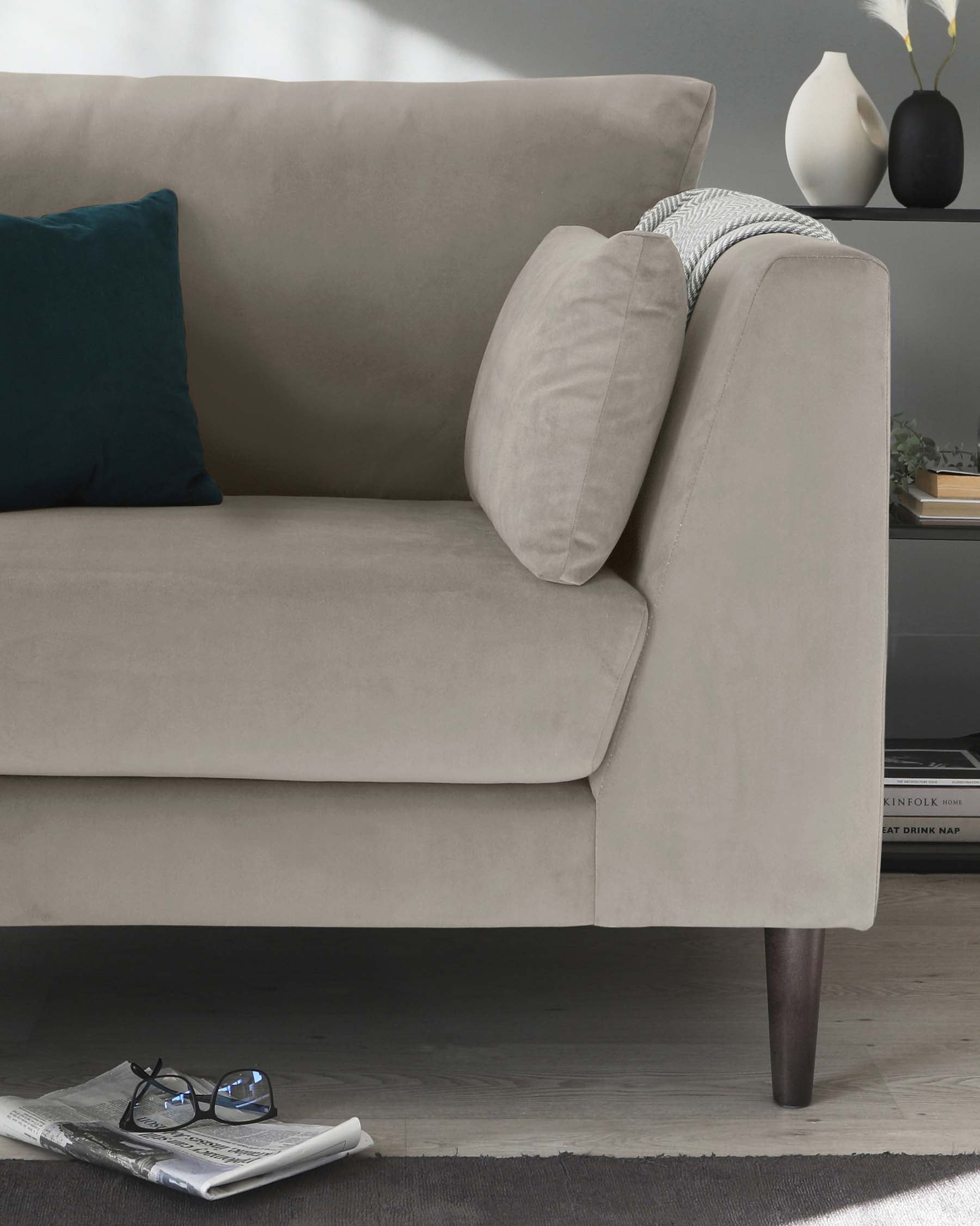 Modern taupe upholstered sofa with dark wooden tapered legs, featuring a smooth fabric finish and including additional comfort with one dark teal and one matching taupe throw pillow. The sofa is accented with a knit grey blanket draped over one arm and set against a deep grey wall, complemented by a simple white vase on a shelving unit in the background. A newspaper and a pair of glasses rest on the dark wood flooring beside the couch, enhancing the cosy and sophisticated atmosphere.