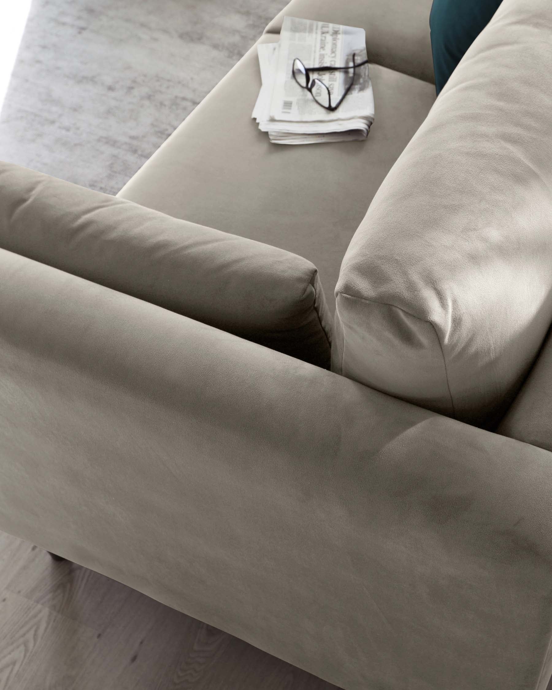 Close-up view of a modern, taupe upholstered sofa with a smooth fabric finish, featuring clean lines, a thick cushioned seat, a straight back, and plush, cylindrical side arms. On the sofa is an unfolded newspaper and a pair of black-rimmed eyeglasses.