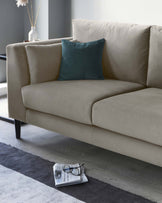 A contemporary beige sofa with a plush finish and clean lines, standing on sleek, dark legs. A single teal accent pillow adds a pop of colour against the neutral upholstery. A low-profile dark grey area rug anchors the space beneath the sofa, with a newspaper and black-rimmed glasses resting on it for a lived-in feel.