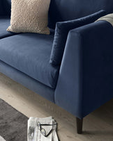 A close-up of a section of a modern navy blue fabric sofa with stylish, clean lines, featuring plush back cushions and a single accent pillow with a textured beige cover. The sofa has a simple armrest design with subtle tufted detailing and sits on dark, tapered wooden legs. In the foreground, a newspaper and a pair of eyeglasses rest on a light wood flooring beside the sofa, suggesting a cosy and sophisticated living space.