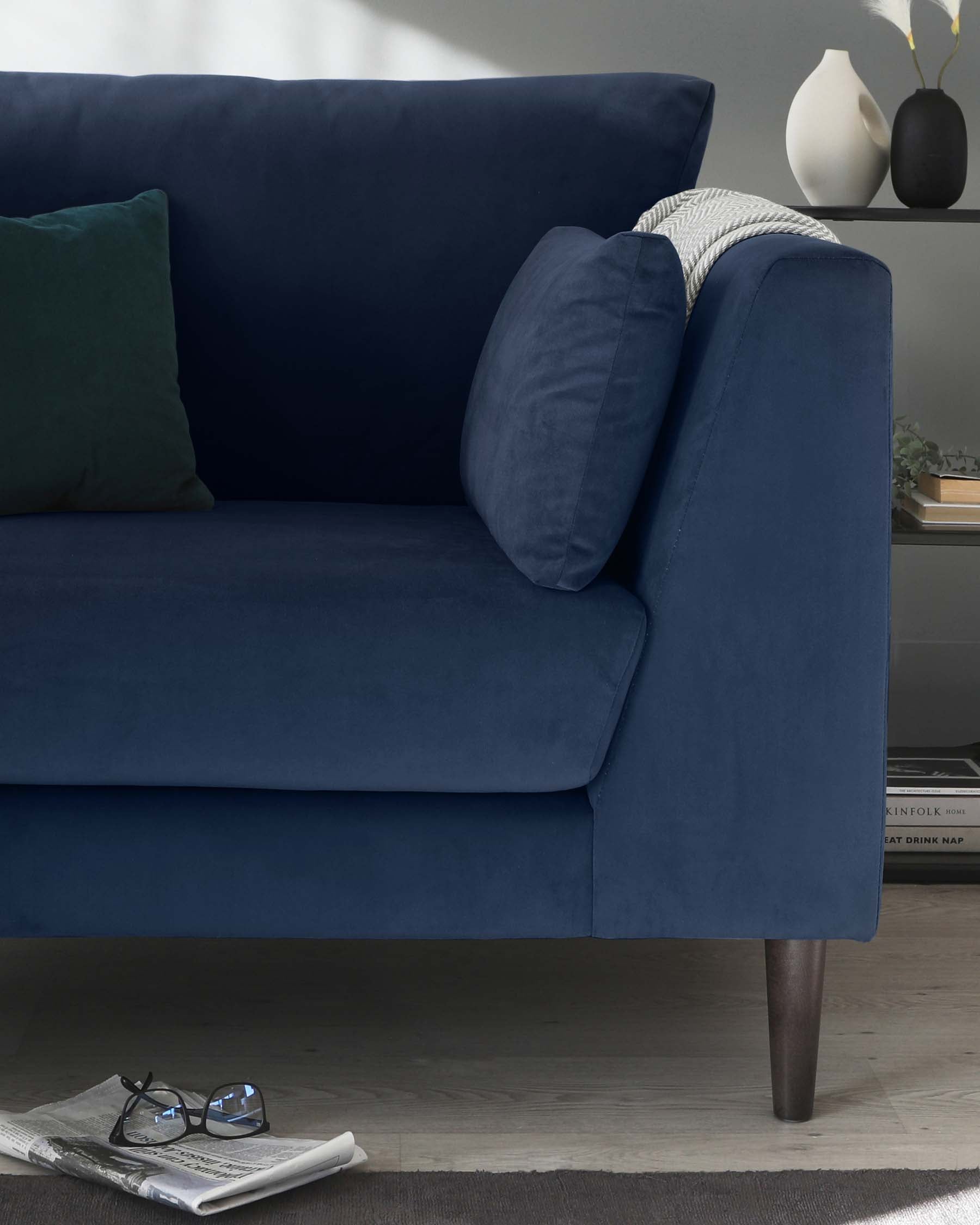 Elegant navy blue fabric sofa with plush cushioning, featuring a single matching bolster pillow and a contrasting green throw pillow. The sofa stands on tapered wooden legs, adding a subtle modern touch to its classic silhouette. A black vase with decorative grass and a side table with books is partially visible in the background, complementing the furniture's staging. On the floor in front of the sofa lies an open newspaper next to a pair of glasses, suggesting a relaxed living space.