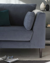 Elegant modern grey fabric sofa with two additional cushions, one teal and one with a geometric pattern, resting against a gently inclined armrest. The sofa is supported by four dark wooden tapered legs, providing a classic touch. A black vase with a white decorative branch is positioned on a shelf in the background, enhancing the contemporary aesthetic, while a casually placed newspaper and glasses rest on the wooden floor beside the sofa, suggesting a lived-in and welcoming space.