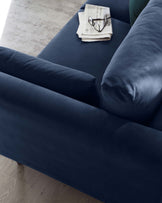 A luxurious navy blue velvet sofa with clean lines and rounded armrests, displayed on a light wooden floor against an abstract-patterned cream rug, accessorized with a casually placed newspaper and reading glasses.