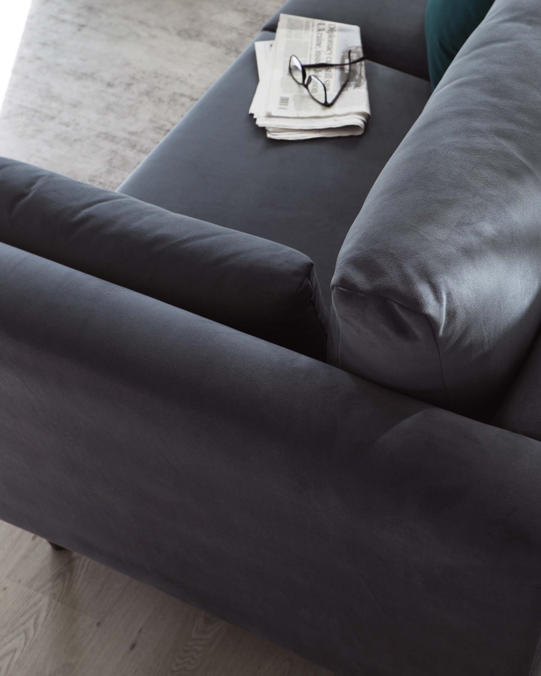 A modern charcoal grey fabric sofa featuring plush back cushions and a sleek silhouette. The close-up angle showcases the soft texture and deep colour of the upholstery, accented by a folded newspaper and a pair of black-rimmed glasses resting on one arm, suggesting a relaxing reading nook. A subtle grey area rug is partially visible, complementing the elegant contemporary aesthetic of the furniture.