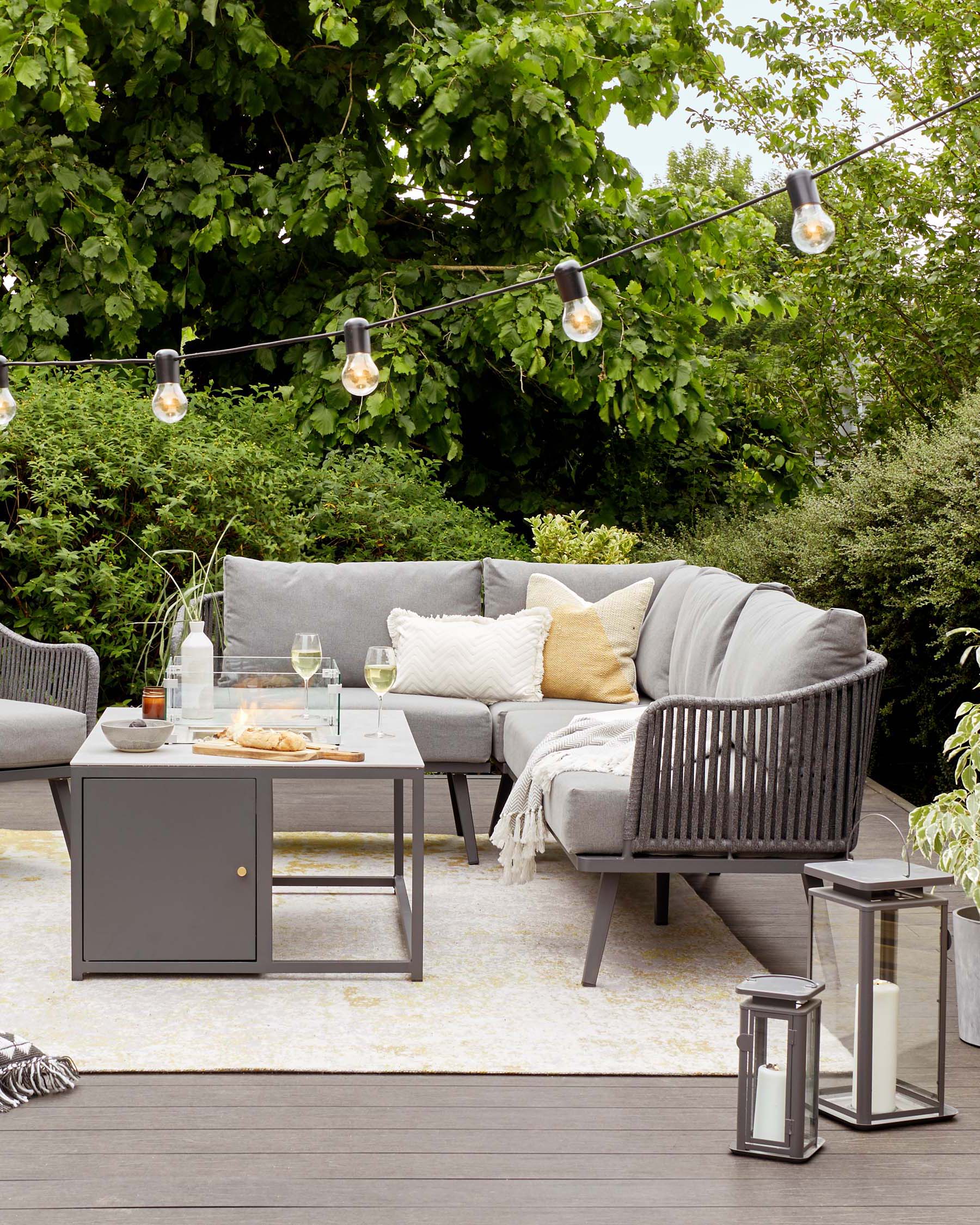 Outdoor furniture set featuring a modern grey sectional sofa with plush cushions, a matching armchair, and a coffee table with dark grey top and storage. Coordinated cushions and throws accent the set. Solar-powered lanterns and festoon lights enhance the ambiance. Set on a wooden deck against a backdrop of lush greenery.