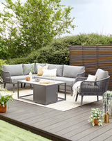 Outdoor contemporary garden furniture set on a wooden deck, featuring a grey sectional corner sofa with plush cushions, a matching armchair, and a rectangular metal coffee table with a simple modern design. The arrangement is complemented by decorative pillows, a light throw, outdoor plants, and ambient lighting accessories, creating a cosy and inviting space for relaxation and entertainment.