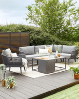 Outdoor patio set featuring a woven grey sofa with matching chairs and plush cushions, complemented by a sleek, rectangular metal coffee table with a glass top. A cosy beige area rug anchors the arrangement on a wooden deck, surrounded by lush greenery.