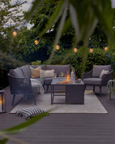 Outdoor patio furniture set displayed on a wood deck, including a modern-style dark grey sofa with plush cushions, two matching armchairs with cushioned seats and vertical slat backs, and a rectangular metal coffee table with a built-in fire pit. All are complemented by decorative pillows, a grey throw, lanterns, and a neutral area rug underfoot, with string lights overhead, creating a cosy evening ambiance.