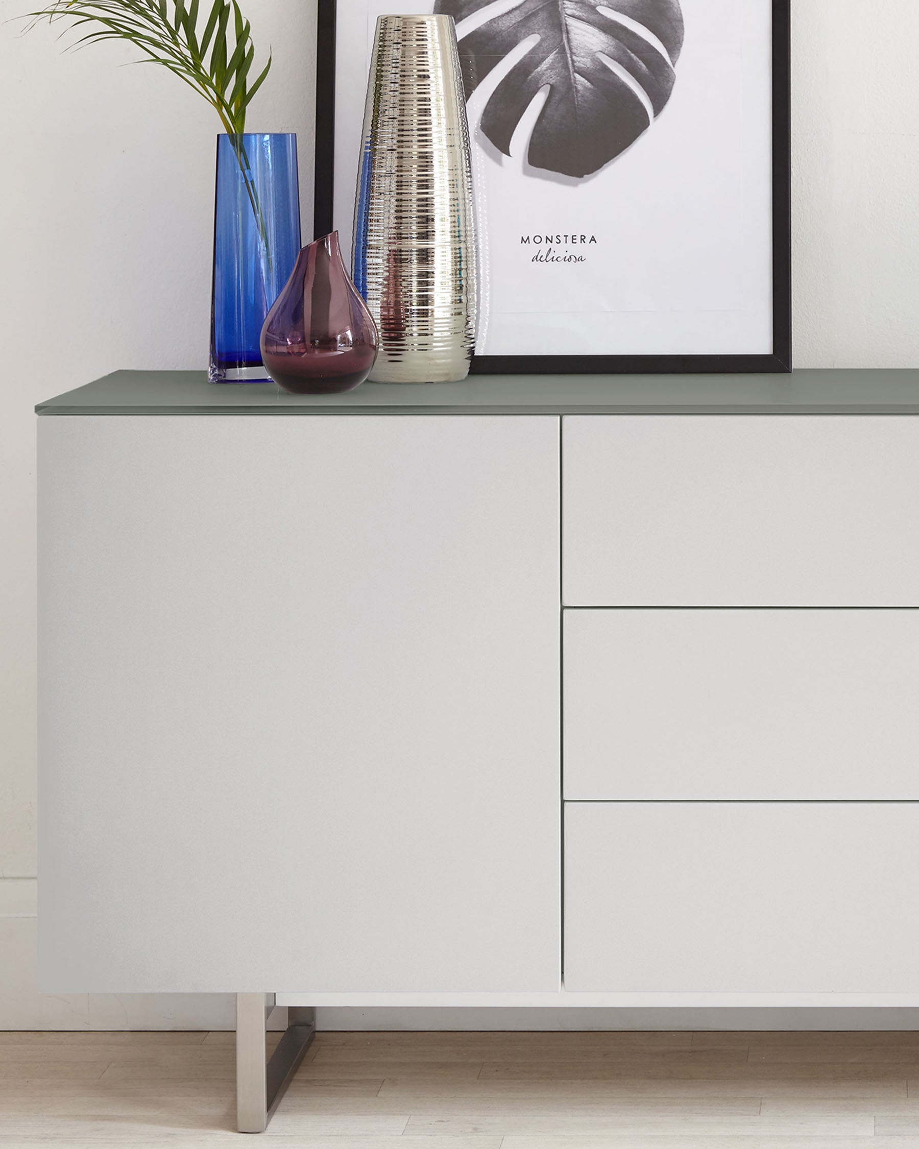 Modern minimalist sideboard in a matte grey finish featuring clean lines, flat-front doors on the left, and three flush drawers on the right with subtle handles. This piece is elevated slightly by a clean, metallic base, offering a sleek, contemporary look suitable for various interior styles.