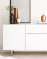 Modern white sideboard with clean lines featuring two cabinet doors and three drawers, standing on sleek metal legs. The surface is styled with decorative items including vases and books.