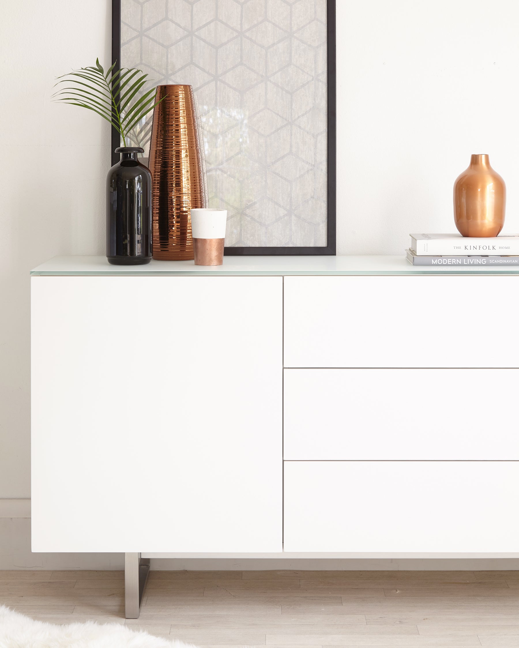 Modern white sideboard with clean lines featuring two cabinet doors and three drawers, standing on sleek metal legs. The surface is styled with decorative items including vases and books.