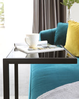 Modern minimalist black metal framed coffee table with a clear glass top, positioned over a plush teal sofa accented with vibrant yellow and teal pillows.