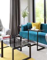 Modern living room set with a plush teal sofa accompanied by a yellow decorative pillow, in front of which sits a sleek glass-top coffee table paired with a smaller nested side table, both featuring black metal frames. The sofa is flanked by a simple, elegant side table holding decorative items.