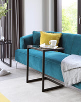 A luxurious teal blue velvet sofa adorned with a mustard yellow pillow with pom-pom trim is paired with a sleek modern side table featuring a minimalist black metal frame and glossy dark tabletop. A complementary light grey fringed throw blanket rests casually on the sofa's arm, enhancing the cosy and stylish ambiance.