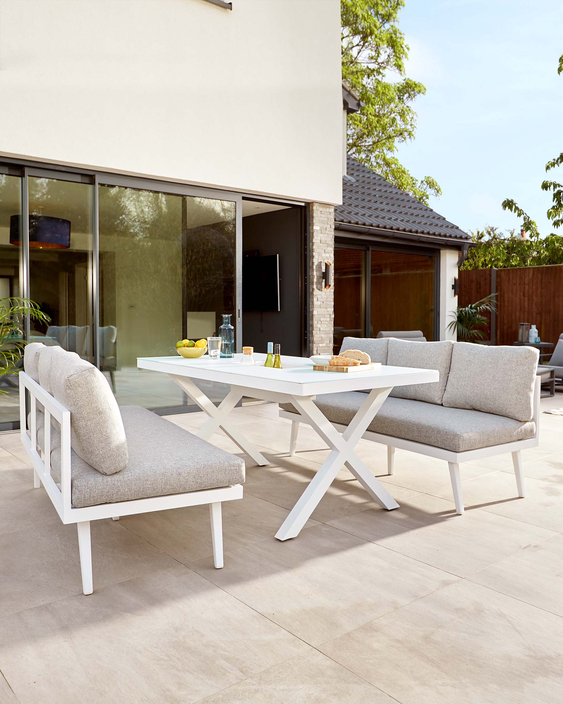 Modern outdoor dining furniture featuring a white rectangular table with a minimalist X-shaped leg design and two matching armless chairs with comfortable grey cushions, arranged on a paved patio.