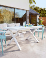 Modern outdoor furniture featuring a white rectangular dining table with a cross-leg design, paired with four aqua blue bistro-style chairs with armrests, set on a patio with a backdrop of sliding glass doors leading to a contemporary home.