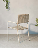 Contemporary minimalist chair with a metal frame and woven seat and backrest, in a neutral colour palette, displayed against a light grey textured wall and floor with a hanging green plant in the background.