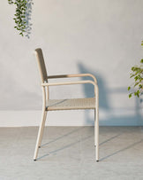 Modern minimalist chair with a beige metal frame and woven seat and backrest, standing on a polished grey floor against a white wall with a hanging green plant in the background.