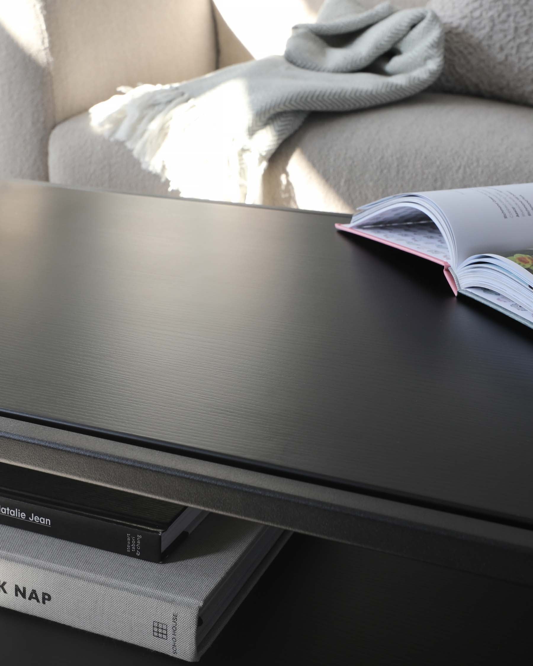 A contemporary coffee table in a modern living room setting, featuring a sleek, dark wood-grain finish and a lower shelf with books. The tabletop is adorned with an open magazine, and behind it, a beige armchair with a soft grey throw blanket is partially visible, suggesting a cosy reading nook.