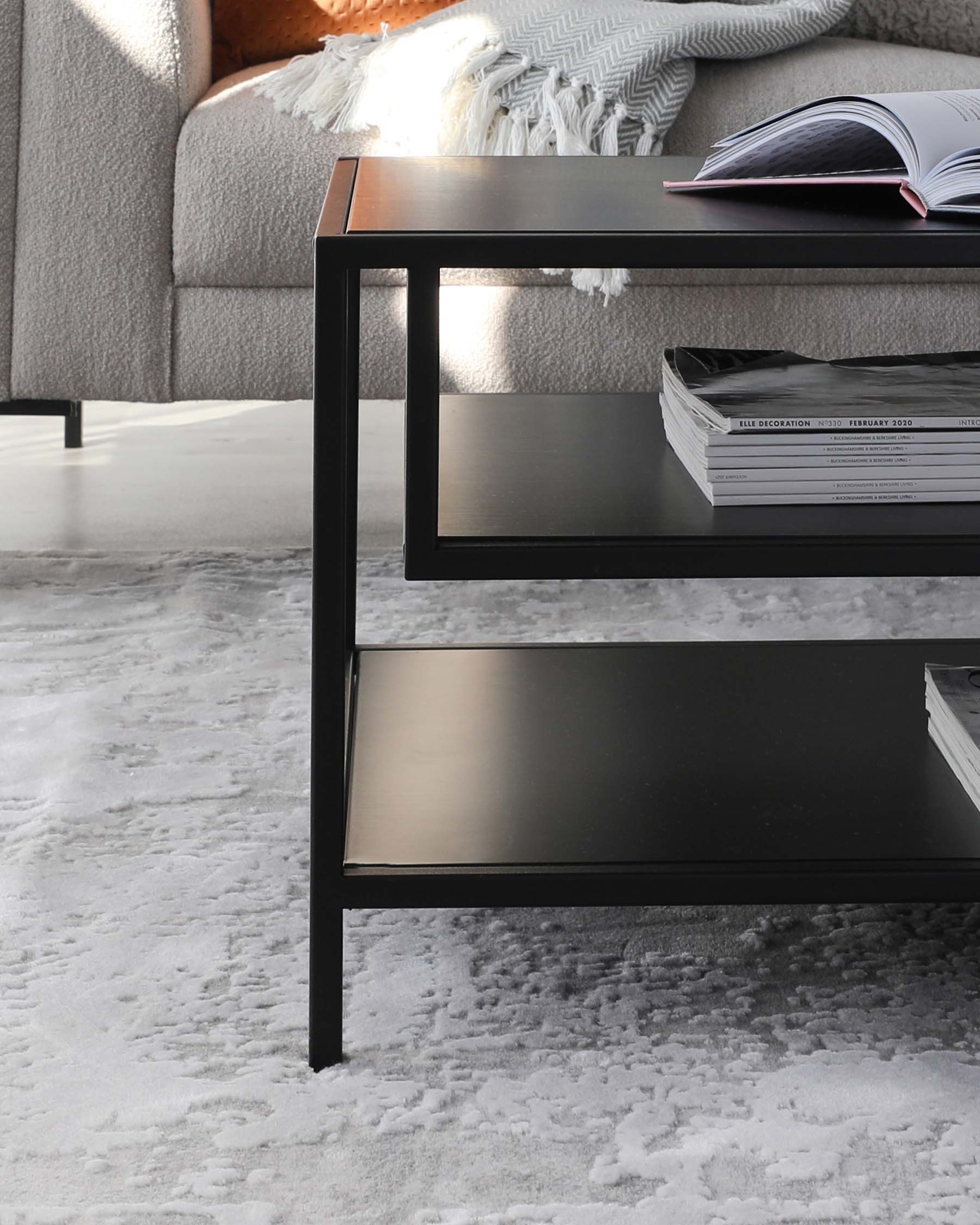 Modern black metal side table with a sleek rectangular shape, featuring two tiers for ample storage, set on a plush grey and white patterned area rug beside a grey upholstered couch with a cosy throw blanket.