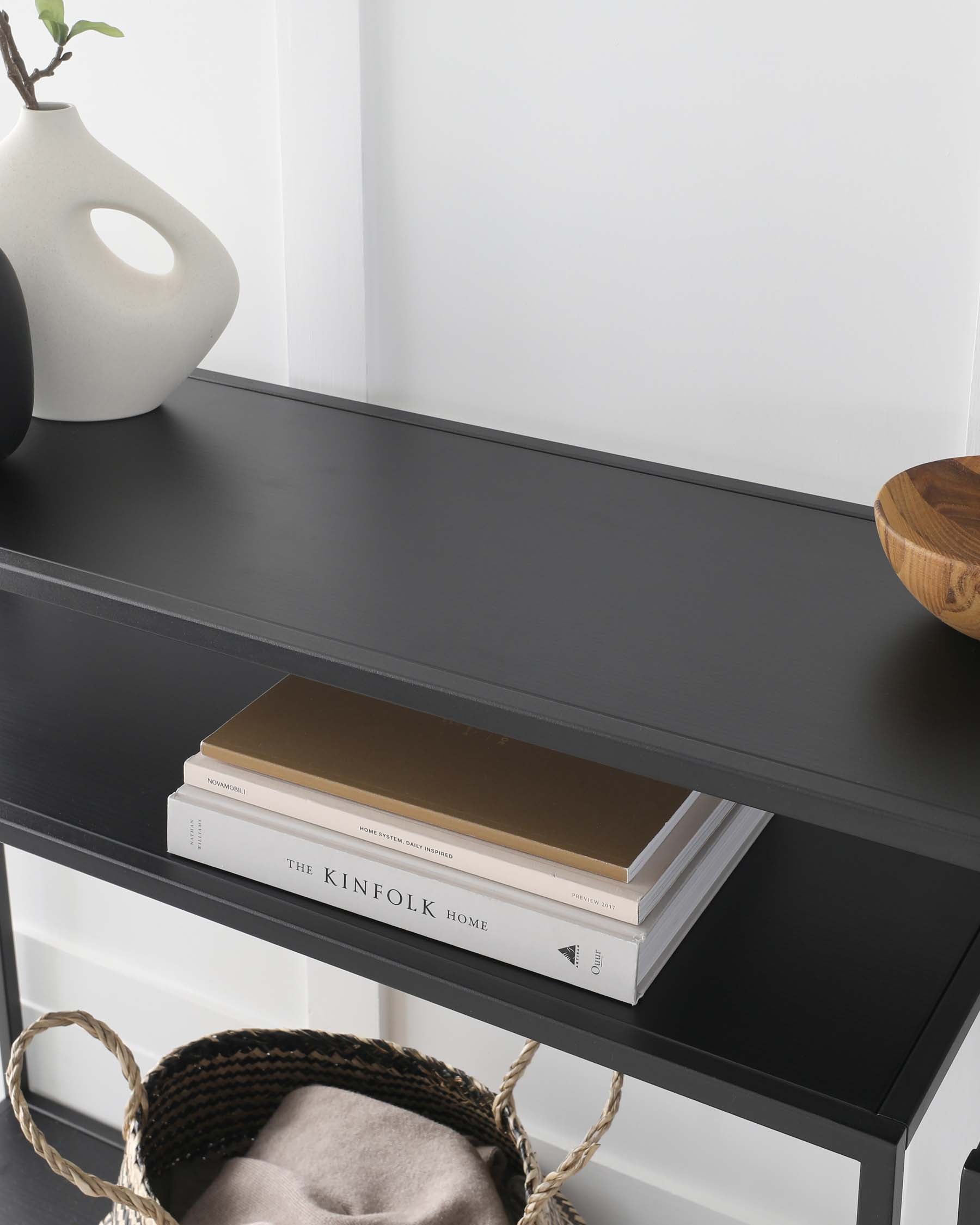 A sleek black console table featuring two tiers, with the top shelf adorned by a uniquely shaped white vase and a wooden bowl, and the lower shelf holding neatly stacked books alongside a woven basket with soft textiles.