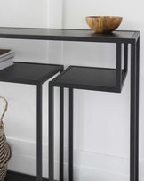 Modern minimalist black console table with a matching pair of nested side tables, featuring clean lines and a sleek finish. The console table has a rectangular top with ample space adorned with a wooden bowl, and it is supported by straight, sturdy legs. The smaller side tables fit neatly under the main table and share the same design aesthetic. A striped woven basket adds a textured accent at the lower side, partially visible.