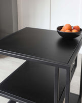 Modern black side table with a matte finish and a lower shelf, featuring metal frame supports and a decorative bowl with oranges on top.