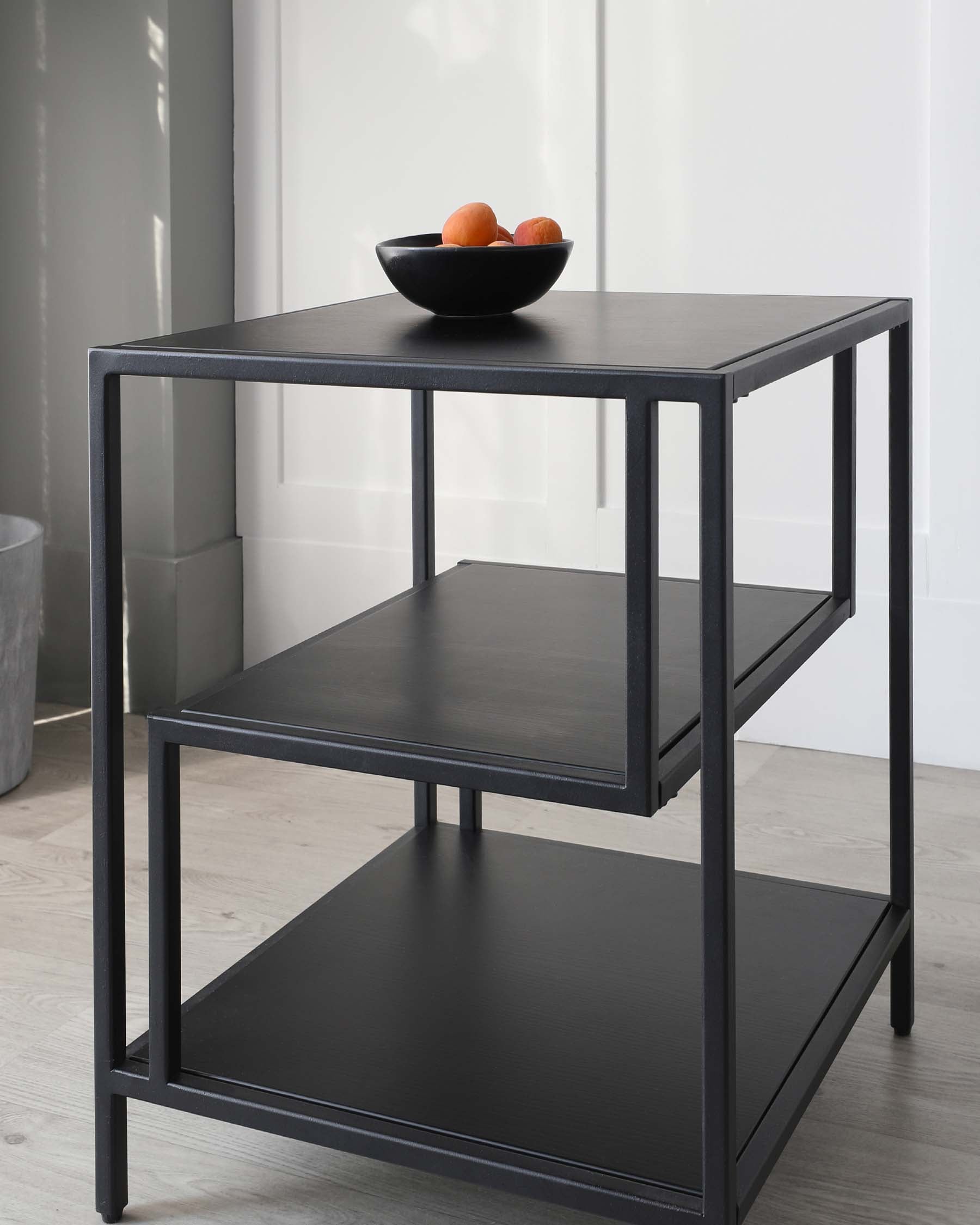 A contemporary, black, two-tiered square end table with a matte finish, featuring straight lines and a minimalist design. The top shelf is adorned with a black bowl containing oranges, and the table is set against a neutral background with light wooden flooring.