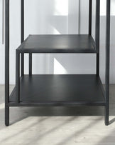 Modern, two-tiered black metal side table with a matte finish. The sleek, square design features clean lines, and each shelf offers ample surface space. Positioned against a white wall on a light wooden floor, highlighting its minimalist aesthetic.