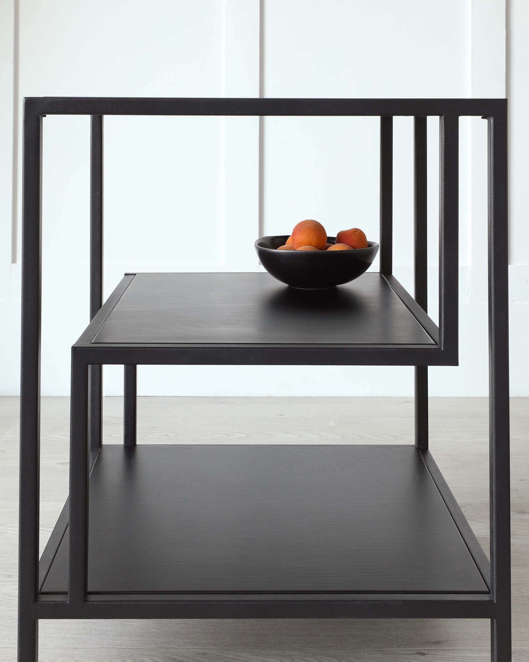 Modern two-tiered square side table in a matte black finish with a simple, minimalist frame and a bowl with oranges on the upper shelf, set against a white background.